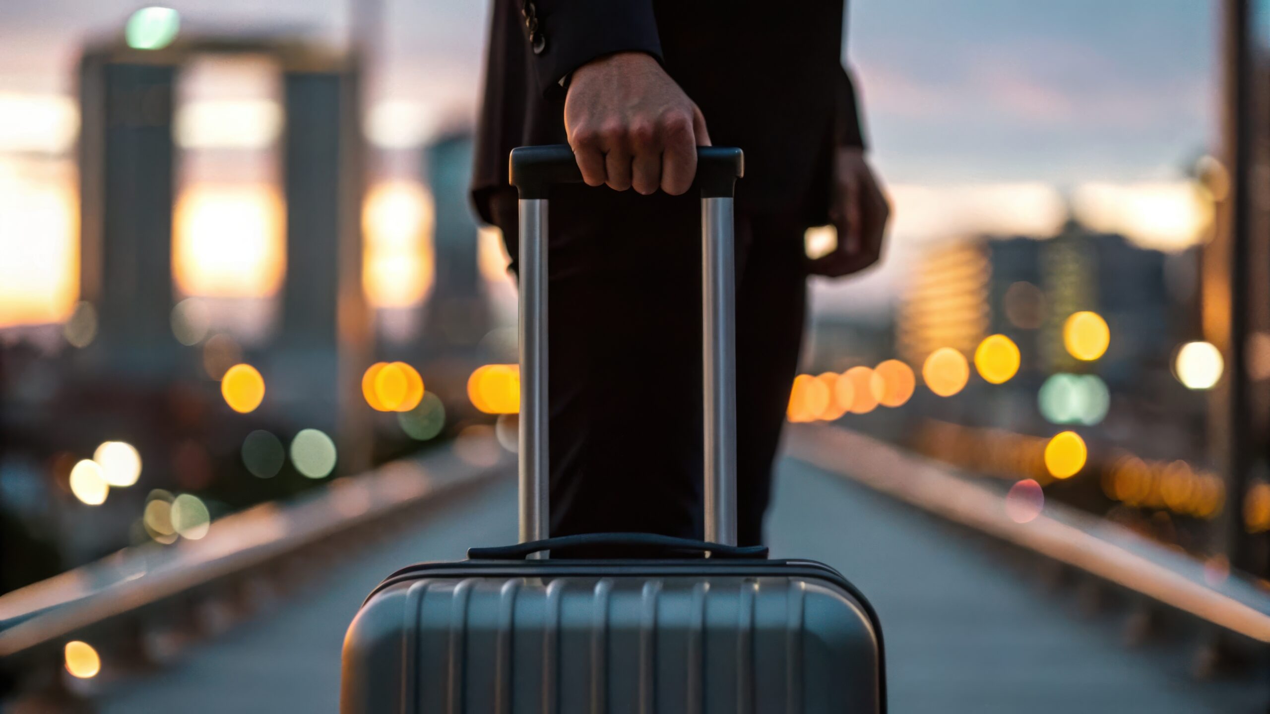 Business travel and global mobility transportation concept. A person holds a suitcase while walking on a city street at sunset, with bokeh lights in the background